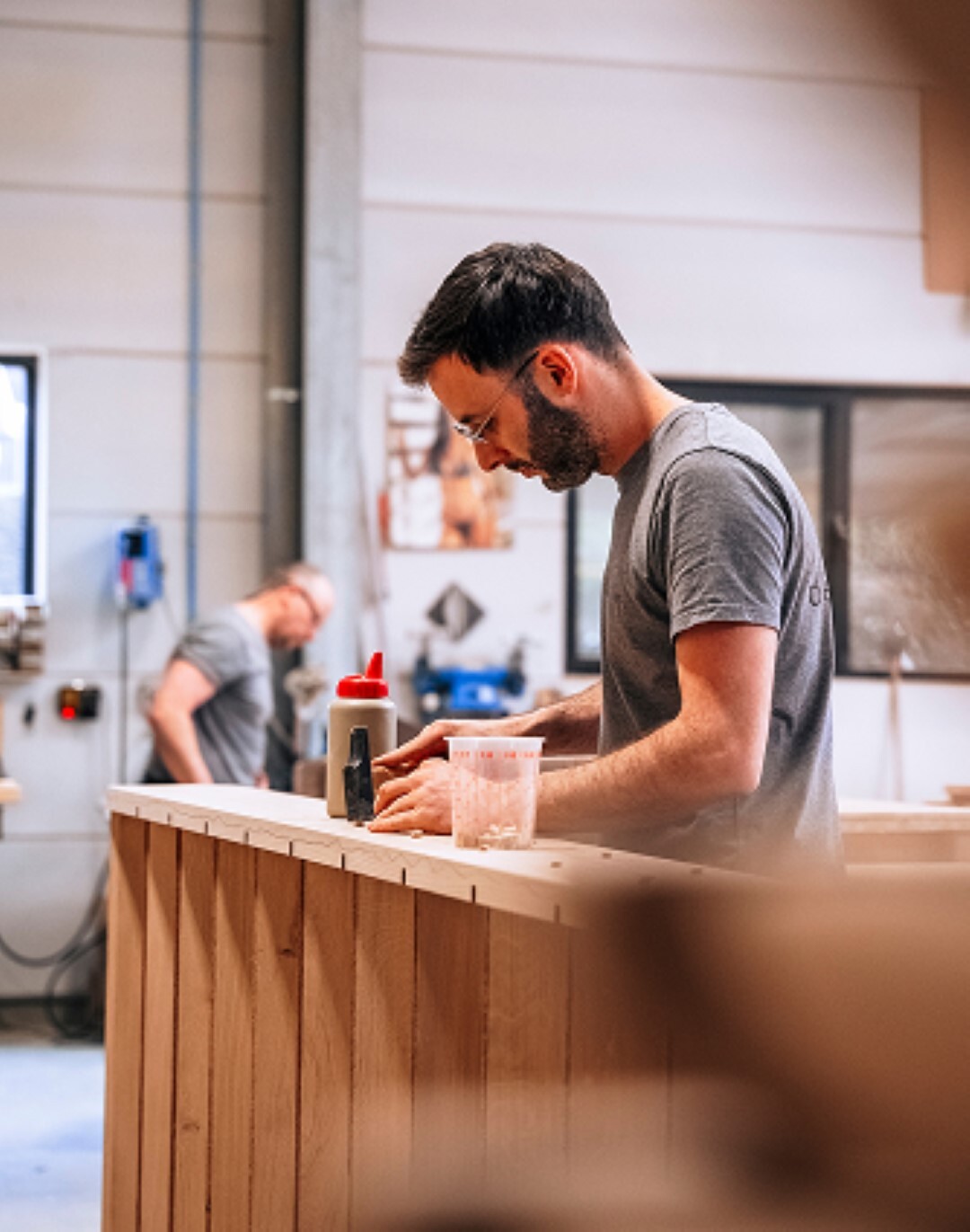 Een man met veiligheidsbril en grijs T-shirt is geconcentreerd aan het werk aan een houten meubelstuk in een goed georganiseerde werkplaats; op de achtergrond is een collega bezig en zijn diverse gereedschappen en materialen zichtbaar.