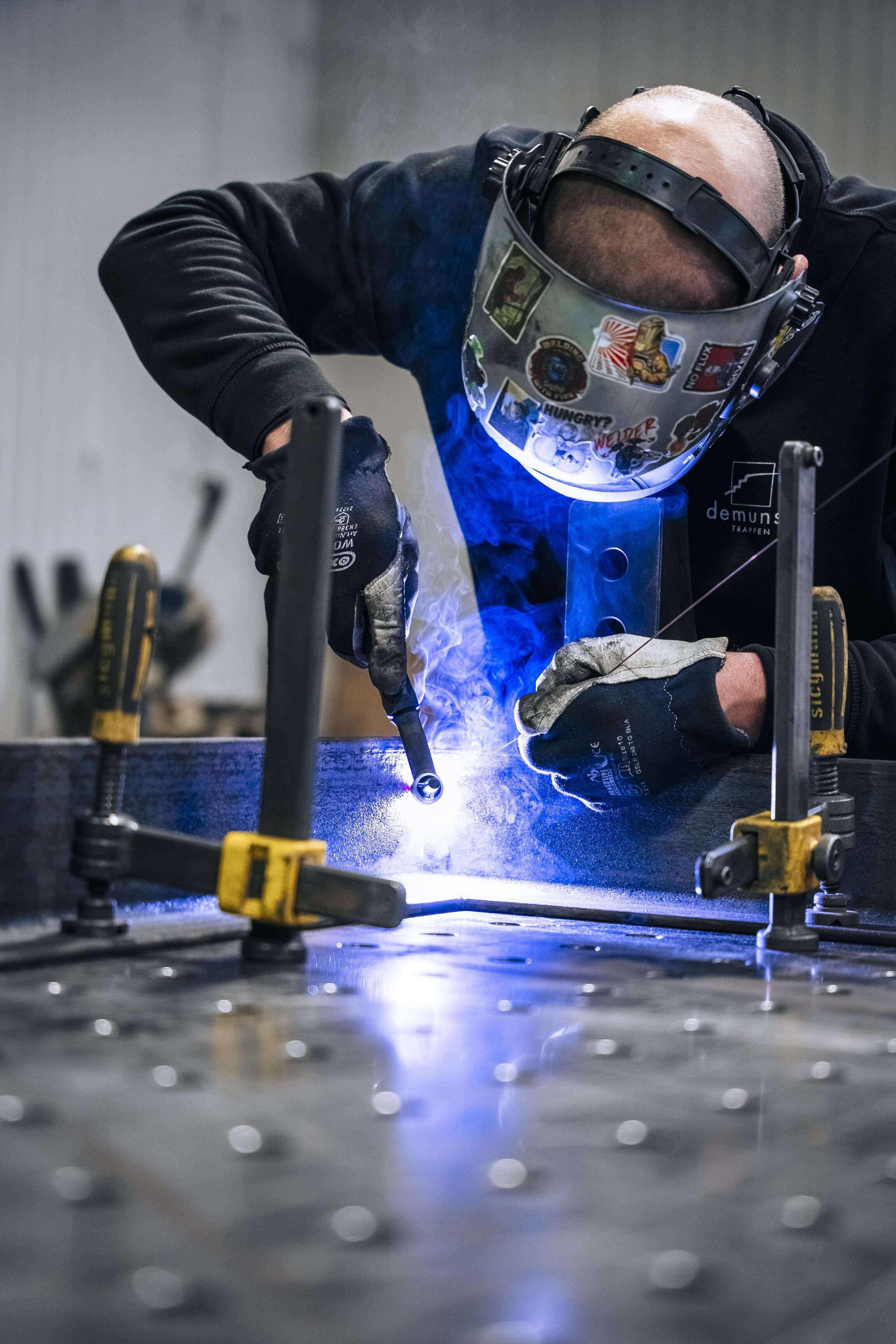 Een man met veiligheidsmasker en handschoenen voert laswerk uit aan een metalen constructie, waarbij fel blauw licht van het lasproces opvalt; de omgeving is een werkplaats met gereedschap op de achtergrond.
