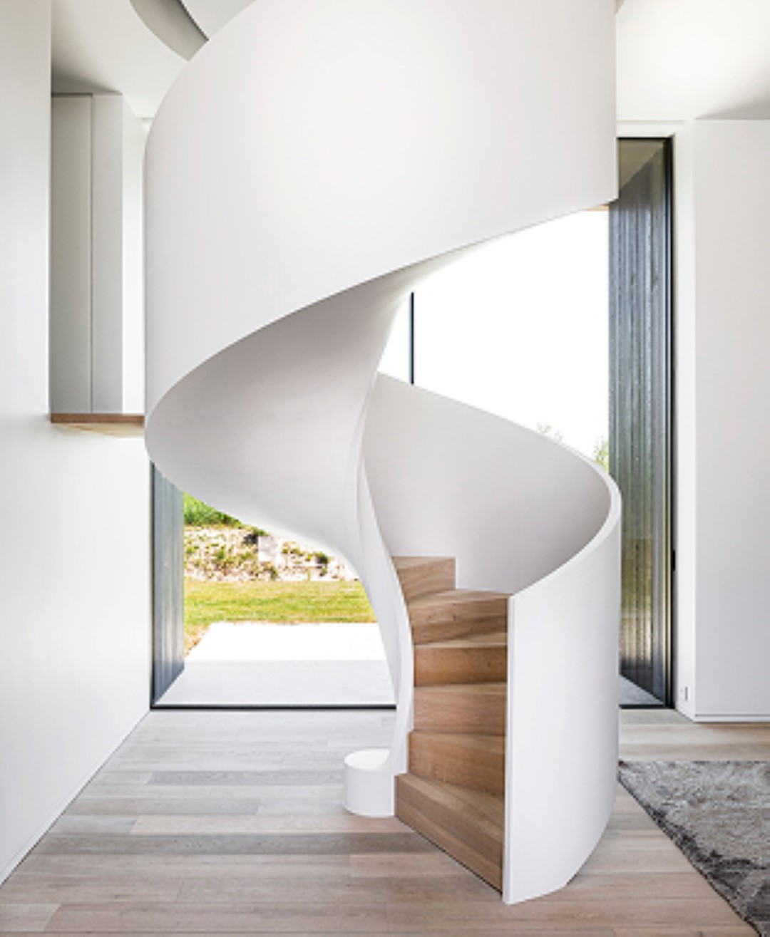 Elegante spiraaltrap met witte, gesloten zijwanden en houten treden, geplaatst in een moderne, lichte ruimte met grote ramen en houten vloer.