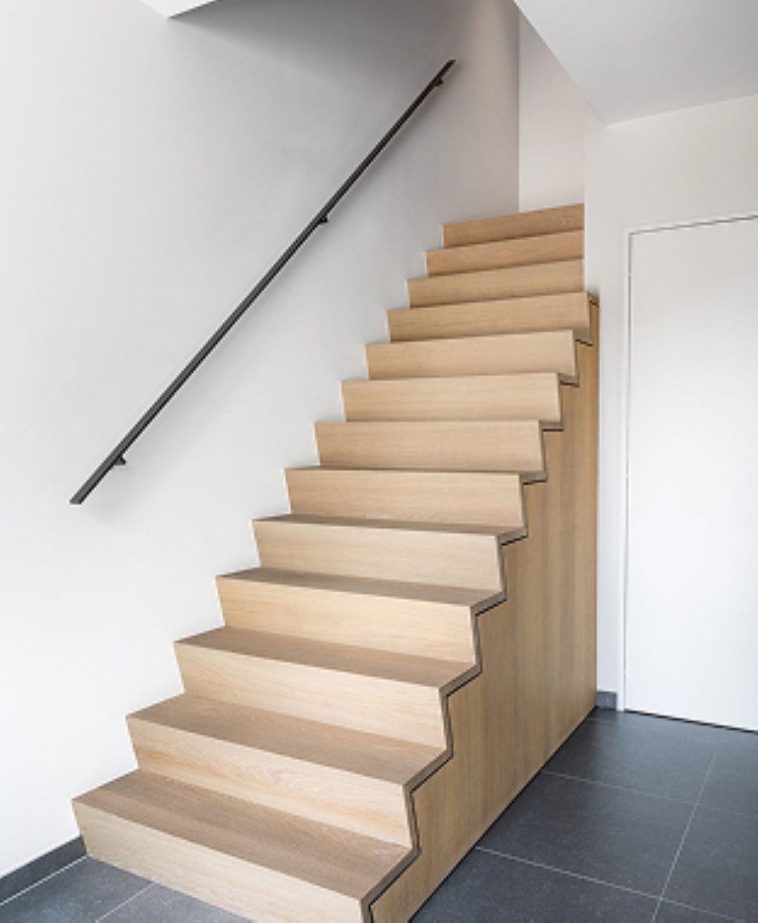 Moderne trap met rechte houten treden in blokvorm, afgewerkt met een zwarte muurleuning, geplaatst in een ruimte met grijze tegelvloer en witte wanden.